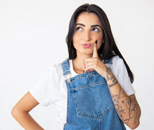 A thoughtful woman with a tattoo on her arm, wearing a white t-shirt and blue overalls.