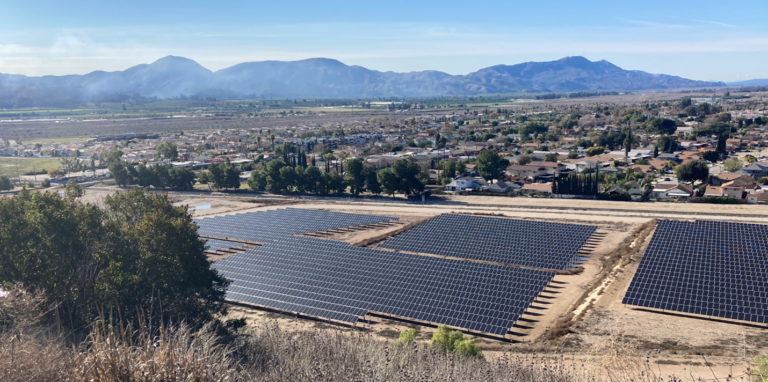 A panoramic view of a solar farm in the foreground with a residential area behind and mountains in the distance under a clear sky.