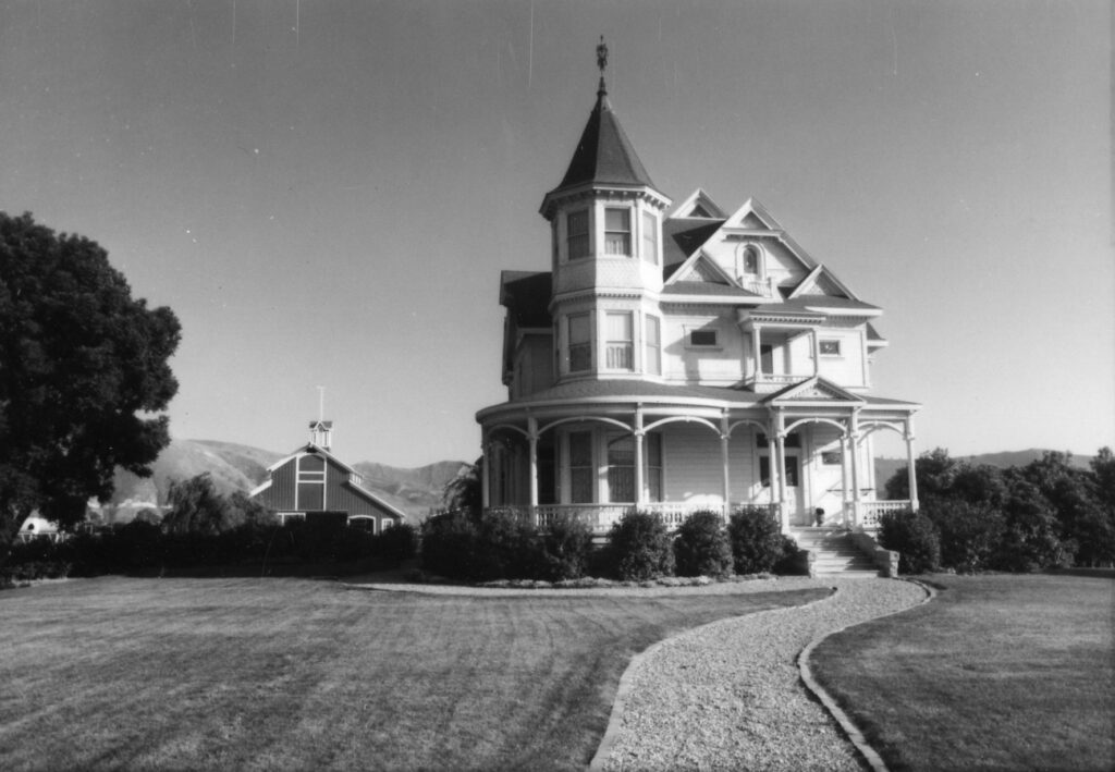 Black and white image of a historic Victorian-style house with a turret and wrap-around porch, situated in a well-maintained garden with a winding gravel path