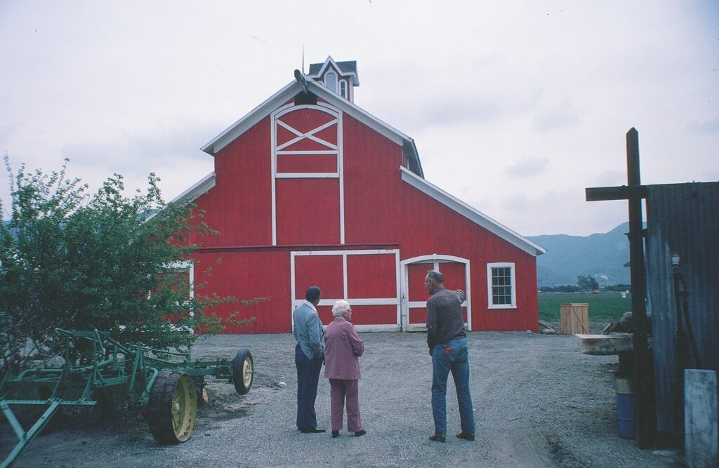 Three adults standing outside a large red barn with farming equipment on a cloudy day.