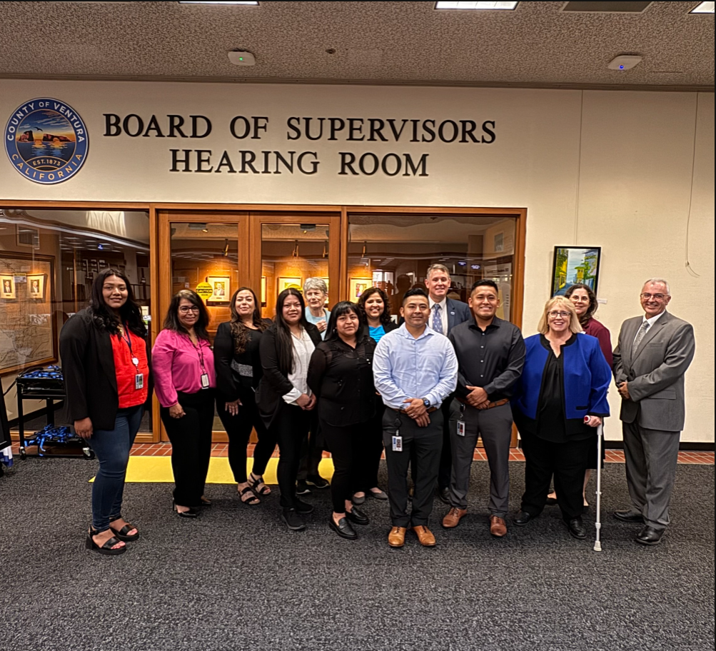 A group of individuals standing together in front of the entrance to the Board of Supervisors Hearing Room, located in Ventura County, California. The sign above the entrance displays the name of the room.