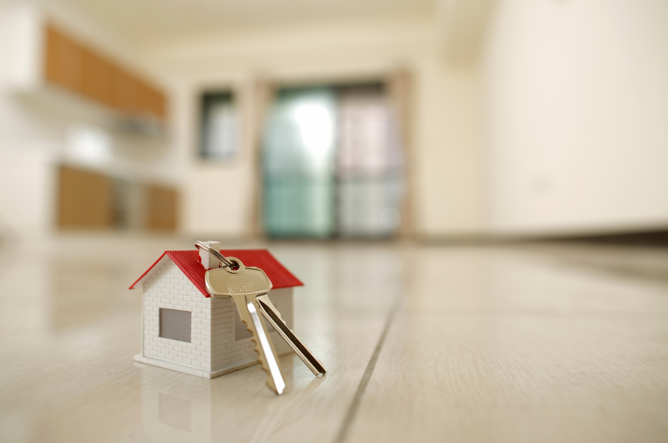 A set of house keys placed on a small model of a house with a red roof, on the floor of an empty, modern living space in the background.