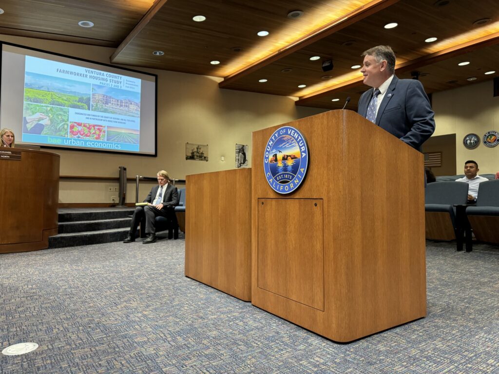 A local Ventura County official speaking at a podium during a government meeting. A presentation slide on 'Ventura County Farmworker Housing Study' is displayed in the background. Another person is seated and taking notes.