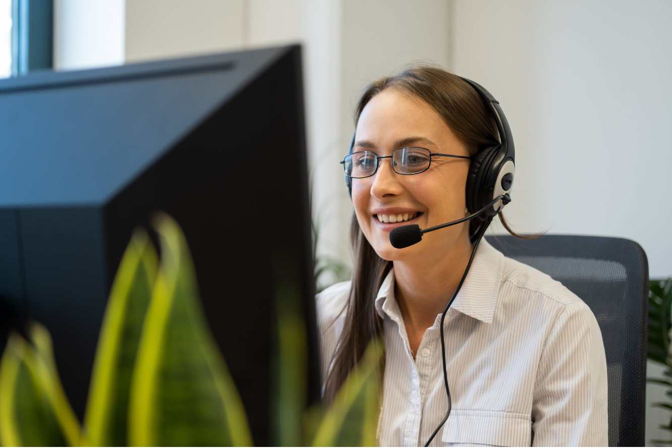 Woman wearing a headset and sitting at a desk, looking at her computer monitor while smiling.