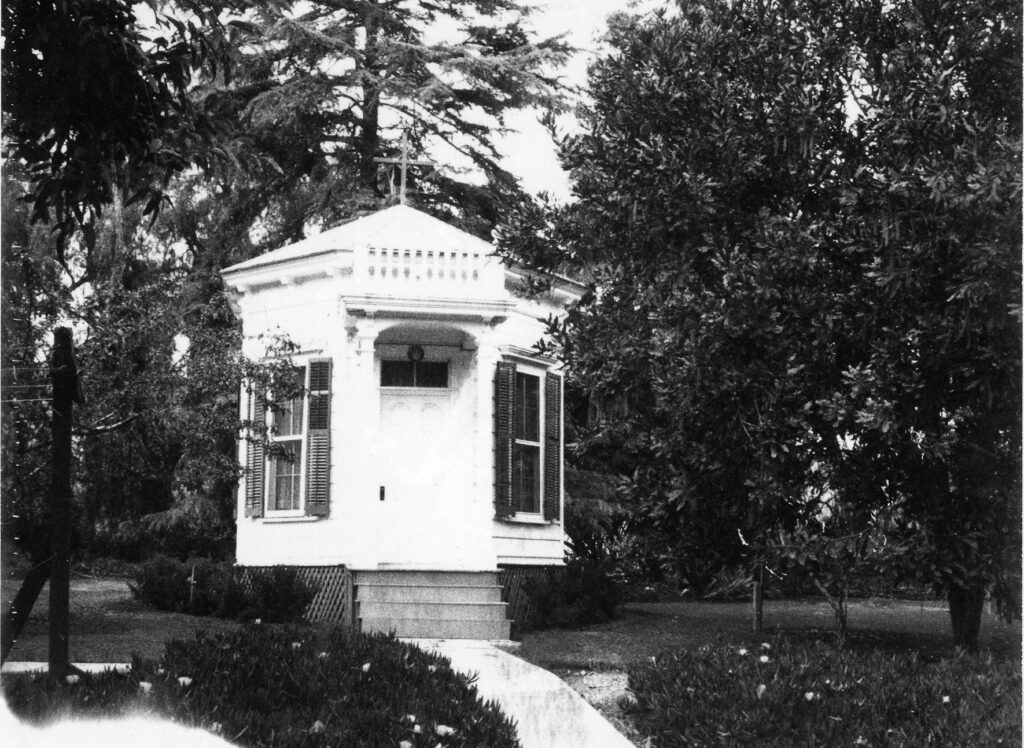 Black and white photo of a small square chapel building with steps and surrounding trees.