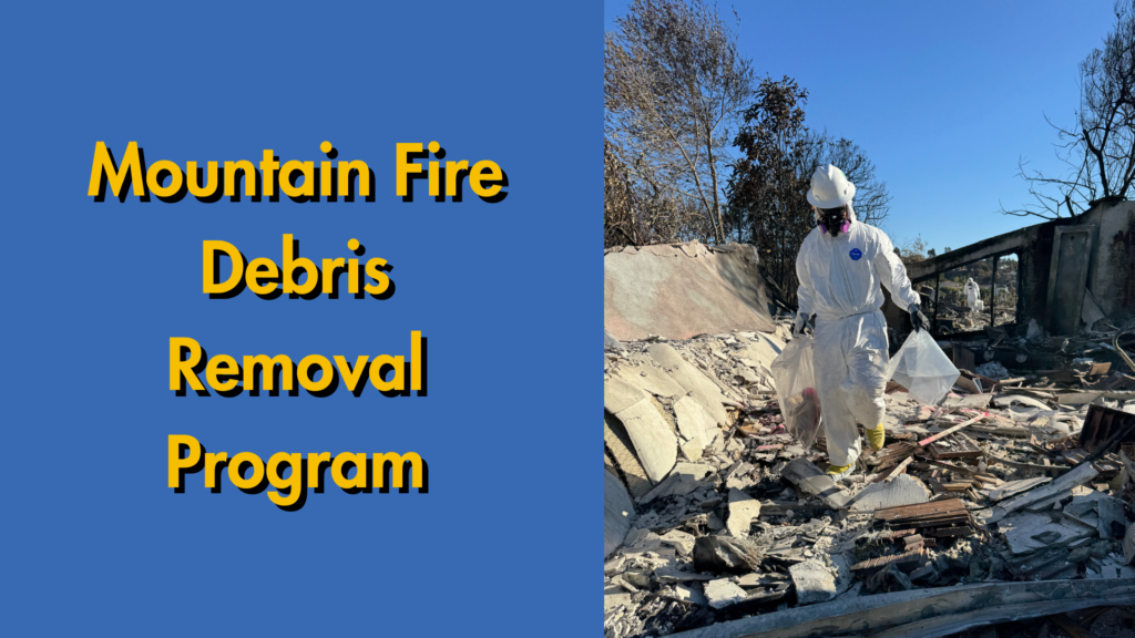 Worker in protective gear removing debris at the site of a burned structure with trees nearby.