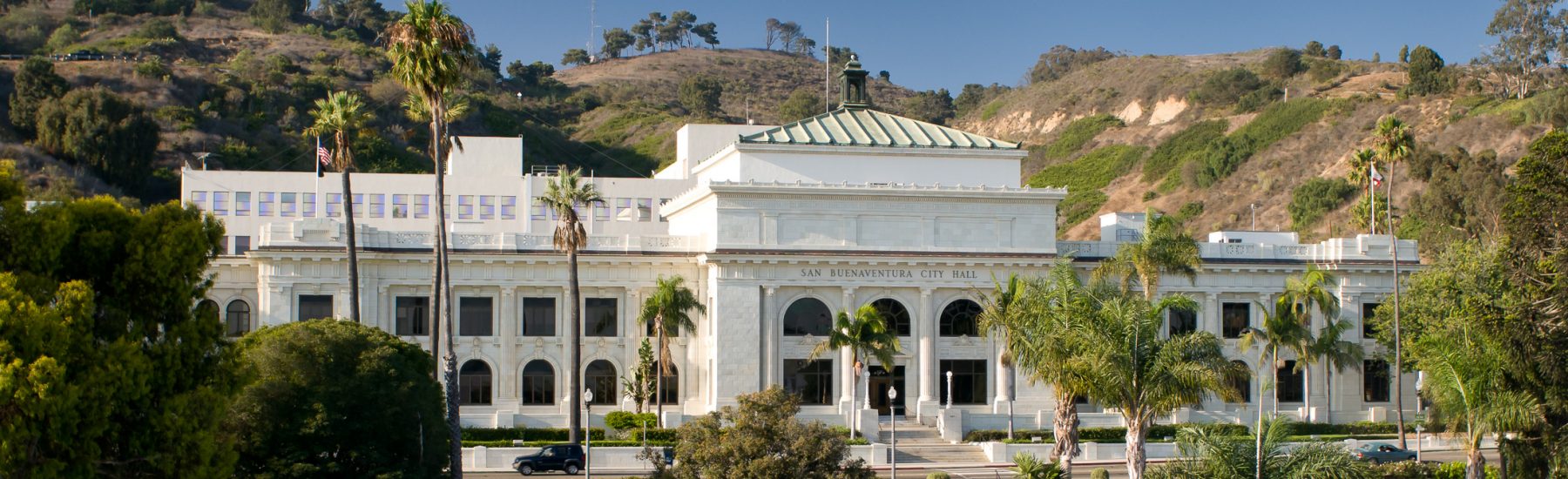 Daylight view of the former Ventura County Courthouse, now San Buenaventura City Hall, with surrounding palm trees and hills.